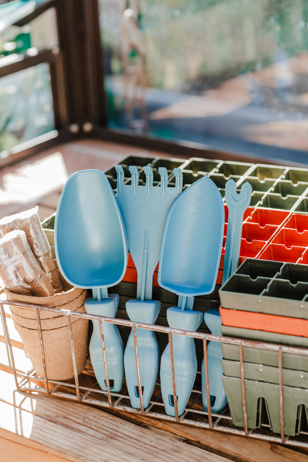 The four Rutabaga Garden Tools, a scoop, weed remover, hand rake, and trowel, stored in a wire basket next to seed starting pots. The blue colored tools are made from recycled end of life marine rope.