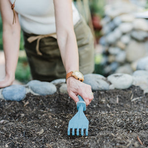 A woman using the hand rake to dig into a garden bed of soil.