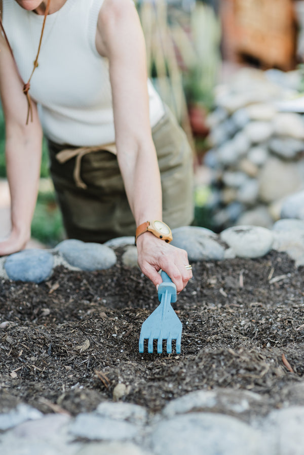 A woman using a blue Rutabaga Garden Tool Hand Rake in the soil of a stacked rock garden bed.