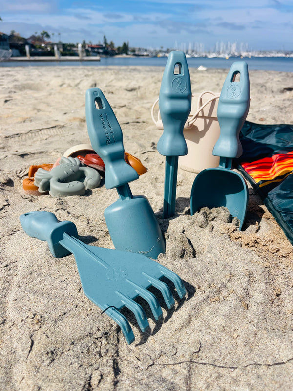 The four Rutabaga Garden tools on the sand. There is a beach blanket and a bucket on one side. On the other side are some sand tools that look like sea creatures.
