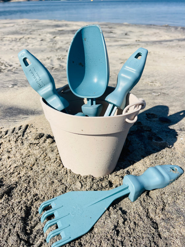 Three Rutabaga Garden Tools are in a white beach bucket. A hand rake is on the sand in front of the bucket.