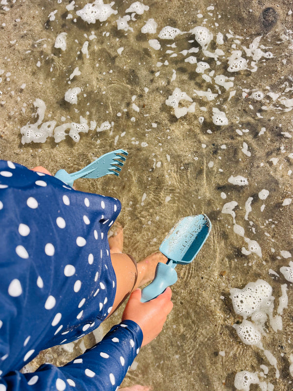 A young child in a polka dot swim shirt wading in the ocean water while holding a blue trowel and hand rake made by Rutabaga Garden Tools.