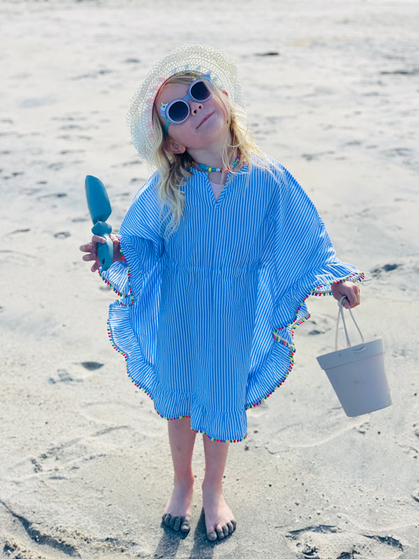 A young girl at the beach in a hat and sunglasses and summer dress. She has a Rutabaga Garden Tool trowel in one hand and a beach bucket in the other hand.