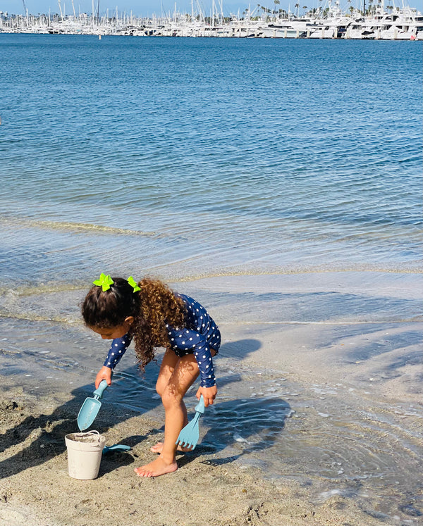 A young girl in a polka dot swim suit that is filling a bucket of sand with a Rutabaga Garden Tool trowel and in her other hand is a Rutabaga Garden Tool hand rake.